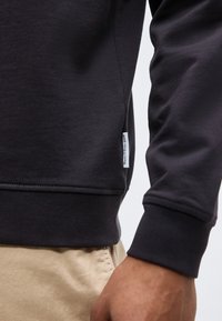 Κοντινό πλάνο ενός ατόμου που φοράει μαύρο φούτερ με μακριά μανίκια, με λευκή ετικέτα στο πλάι και μπεζ παντελόνι, εμφανίζοντας το κάτω μέρος του κορμού και το χέρι.