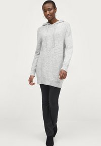 Hellgraues Kapuzenkleid aus weichem Material mit langen Ärmeln, Kordelzugkapuze und seitlichen Schlitzen, kombiniert mit schwarzen, eng anliegenden Hosen.