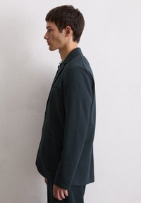 Donkerblauwe blazer van gestructureerde stof. Heeft een reverskraag, een enkele borstzak en twee voorzakken. Rechte silhouet.