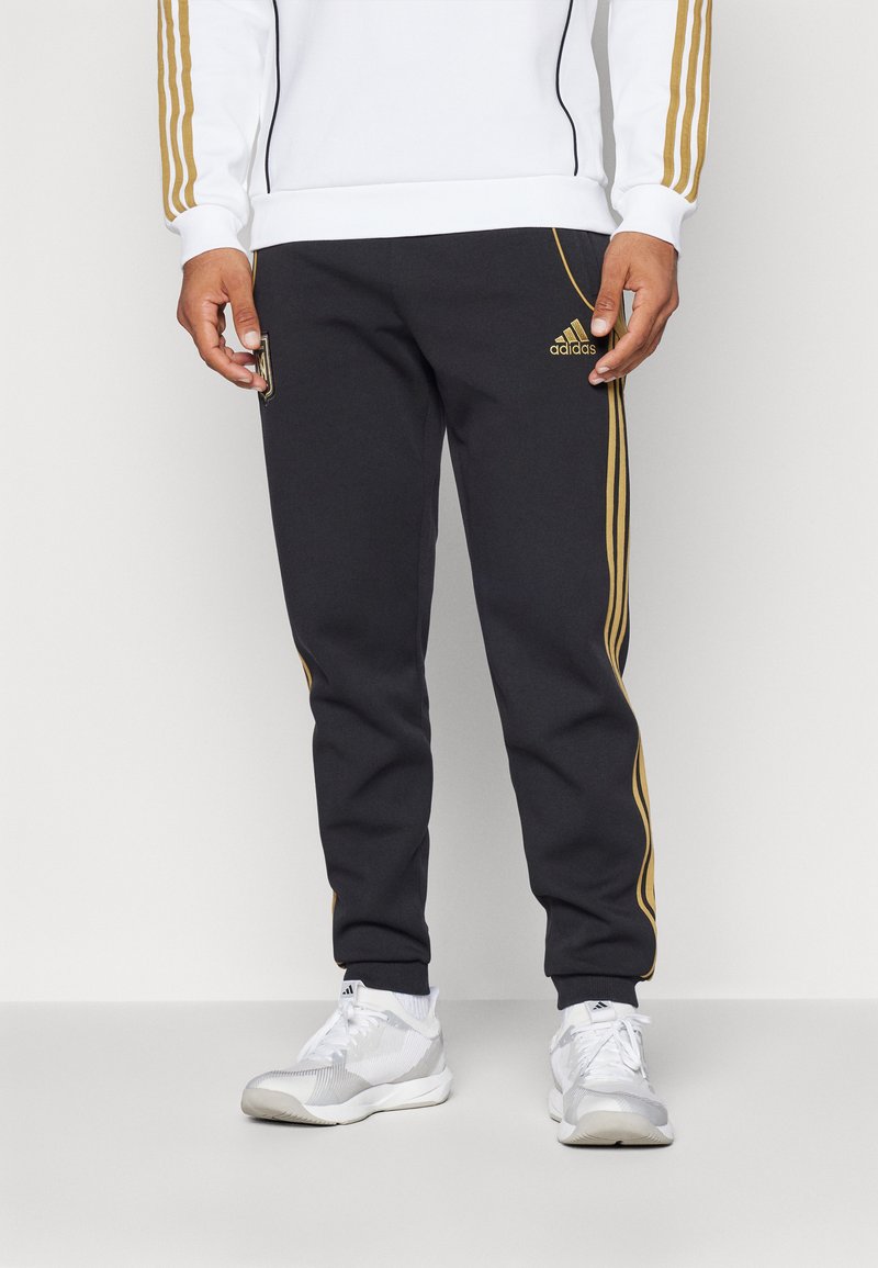 Pantalons de sport noirs avec des bandes latérales dorées, un logo et des poignets aux chevilles. Associés à des baskets gris clair avec un motif texturé.