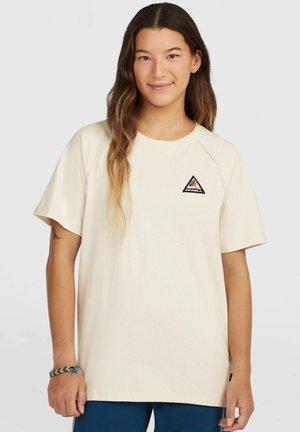Młoda osoba z długimi brązowymi włosami, ubrana w beżowy T-shirt z małym trójkątnym logo na piersi oraz kolorową plecioną bransoletką.