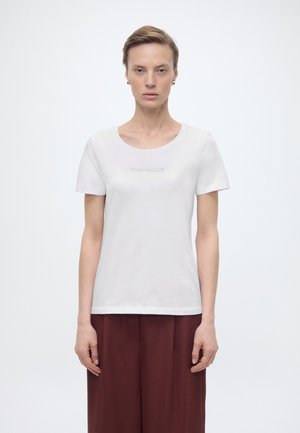 Femeie cu păr scurt purtând cercei aurii, un tricou alb Tom Tailor și pantaloni largi bordo, stând în fața unui fundal alb simplu.