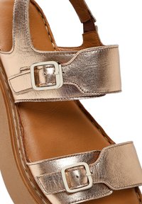 Sandal in metallo dorato con due cinghie regolabili dotate di fibbie quadrate. Il materiale presenta una texture liscia, mentre la soletta è in pelle marrone.