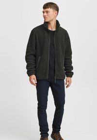 Dark green fleece jacket with a high collar, zip front, and side pockets. Paired with black pants and brown hiking shoes.