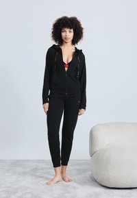 Svart zip-up hoodie och åtsittande byxor med röda accentfärger, kombinerat med en svart sport-bh. Slät tygkvalitet, avslappnad design, modellen står barfota.