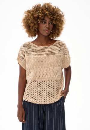 Kobieta o kręconych włosach, ubrana w beżowy, wzorzysty sweter z dzianiny i granatowe spodnie w wąskie paski, stoi z jedną ręką w kieszeni.