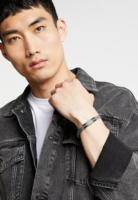 Tummanharmaa farkkutakki, jossa näkyvät saumat; mallilla on vasemmassa ranteessaan metallinen rannekoru, jossa on teksturoitu yksityiskohta. Valkoinen t-paita alla.