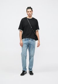 Zwart T-shirt met korte mouwen en witte biezen, lichtblauwe spijkerbroek en zwarte sneakers. De persoon houdt een kleine zwarte schoudertas vast.