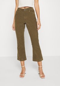 Une femme portant un pantalon en velours côtelé vert olive, un haut sans manches blanc et des sandales à talons blancs, se tenant contre un fond uni.