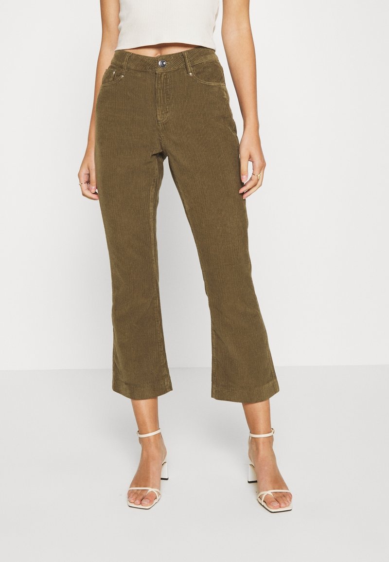 Une femme portant un pantalon en velours côtelé vert olive, un haut sans manches blanc et des sandales à talons blancs, se tenant contre un fond uni.