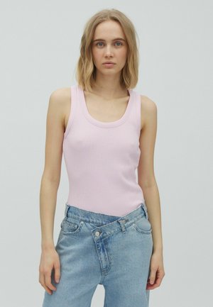 Młoda kobieta z blond włosami do ramion, ubrana w jasnoróżowy top i niebieskie dżinsy z wysokim stanem na jednolitym tle.