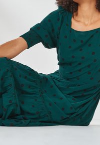 Grünes Kleid aus leichtem Stoff, mit schwarzen Punkten, kurzen Ärmeln, einem quadratischen Ausschnitt und gerafften Details am Saum.