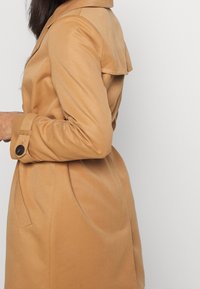 Tepelný trenčkot v béžové barvě s klopovým límcem, dlouhými rukávy s knoflíkovými manžetami a rozšířeným detailem na zadním díle. Hladká, přizpůsobená tkanina.