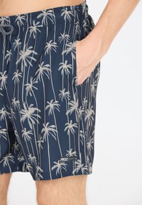 Marinefarbene Schwimmshorts mit Palmenmuster, elastischem Bund, Kordelzug, Seitentasche und einem schwarzen Etikett mit "CRUZ" am Saum.