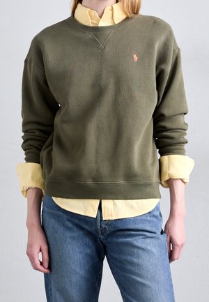 Person iført gul skjorte under olivengrøn sweatshirt med logo, kombineret med blå jeans, stående mod en ensfarvet baggrund.