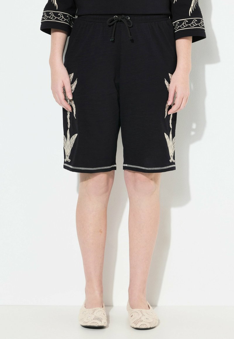 Pantaloni neri con vita regolabile, caratterizzati da ricami floreali crema sui lati e un orlo semplice. Tessuto leggero.