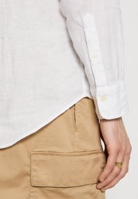 Bras droit en chemise blanche à manches longues et main avec bague en or reposant près de la poche d'un pantalon beige avec rabat.