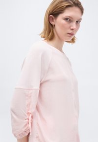 Νέα γυναίκα με κοντά ξανθά μαλλιά που φοράει ένα ανοιχτό ροζ μακρυμάνικο τοπ με μανίκια μαζεμένα και δεμένα με φιόγκους, στραμμένη ελαφρώς πλευρικά.