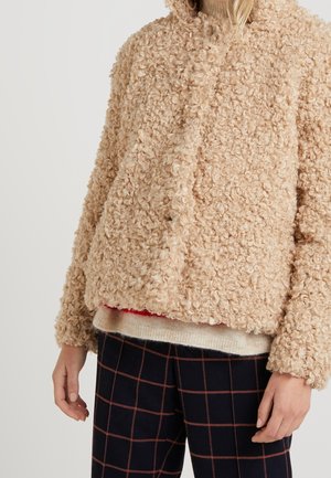 Kvinde iført en beige krøllet tekstureret jakke over en lys sweater og sorte bukser med brunt ternet mønster, hænder afslappet langs siderne.
