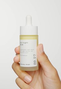 Bouteille en verre clair contenant un sérum jaune, avec un capuchon compte-gouttes blanc. L'étiquette affiche le nom du produit et les ingrédients en texte noir.