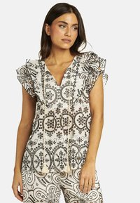 Top blanco con patrones florales negros, mangas con volantes y un escote en V profundo. Presenta borlas decorativas en la parte delantera. Combinado con pantalones estampados.