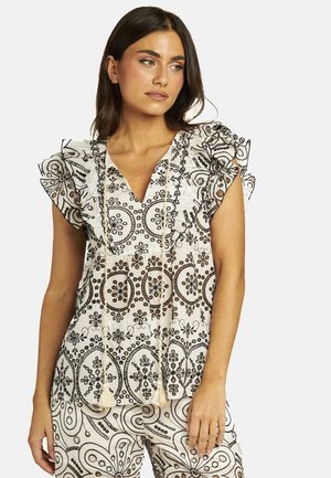 Top blanco con patrones florales negros, mangas con volantes y un escote en V profundo. Presenta borlas decorativas en la parte delantera. Combinado con pantalones estampados.