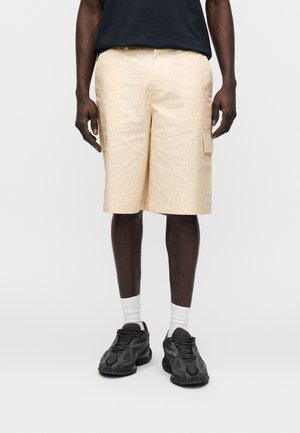 Beige korte broek met een gestructureerd ontwerp, voorzien van zijzakken en een ontspannen pasvorm. Gecombineerd met zwarte sneakers en witte sokken.