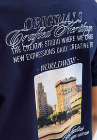 Tamsiai mėlynas medvilninis marškinėliai su baltais siuvinėtais užrašais ir pastatų grafika. Dizainas apima stačiakampį vaizdą ir dekoratyvinius akcentus.