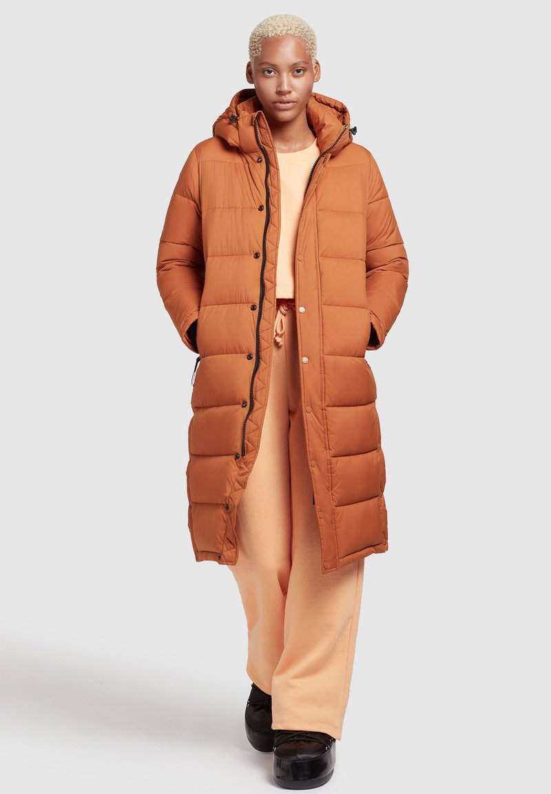 Langer, gepolsterter Mantel in warmem Orange, mit Kapuze, Druckknöpfen und Seitentaschen. Kombiniert mit hellorangefarbenen Hosen und schwarzen Schuhen.