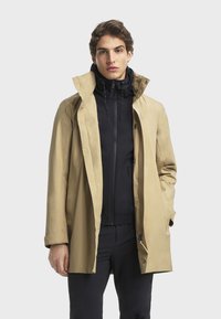 Beige Trenchcoat mit hohem Kragen, über einer taillierten schwarzen Jacke getragen. Verfügt über einen lässigen Schnitt und strukturierte Schultern.