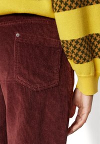 Maroon corduroy pants with vertical ribbing, featuring a back pocket and a metal button detail, paired with a yellow and brown striped sweater.