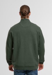 Mann mit braunen Haaren im Dutt, der einen dunkelgrünen Sweatshirt und blaue Jeans trägt, mit dem Rücken zu einer einfarbigen hellen Hintergrund.