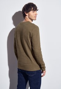 Olivgrön långärmad skjorta i texturerad tyg med rundad hals, kombinerad med mörkblå jeans. Synlig sömnadsdetalj vid fållen.