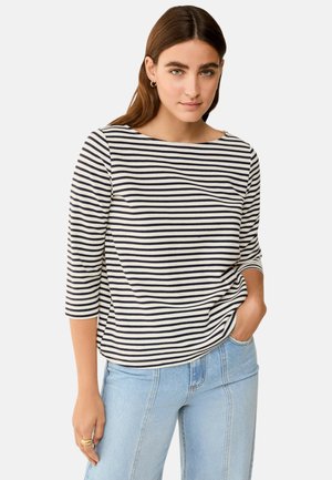 Donna che indossa una maglietta a righe orizzontali bianco e nero con scollo a barca e maniche a tre quarti e jeans azzurri, mano in tasca.