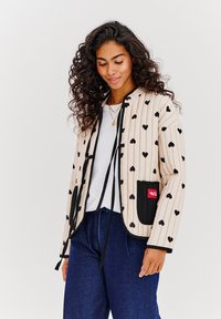 Prešívaná bunda krémovej farby s čiernymi srdiečkovými vzormi, s predným zaväzovaním, čiernymi akcentmi na vreckách a uvoľneným strihom.