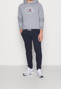 Grauer Kapuzenpullover mit Frontlogo, Kordelzug und Kängurutasche, kombiniert mit dunkelblauen Jogginghosen und weißen Sportschuhen.