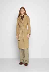 Mantel aus Kamelfell-Wollmischung mit einem taillierten Gürtel, Spitzrevers und zwei vorderen Taschen, kombiniert mit beigen weiten Hosen und braunen Schuhen.