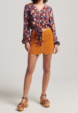 Kvinde iført en blomstret bluse med lange ærmer og bindebånd foran, orange mininederdel med slids foran og brune platåsandaler med ankelremme.