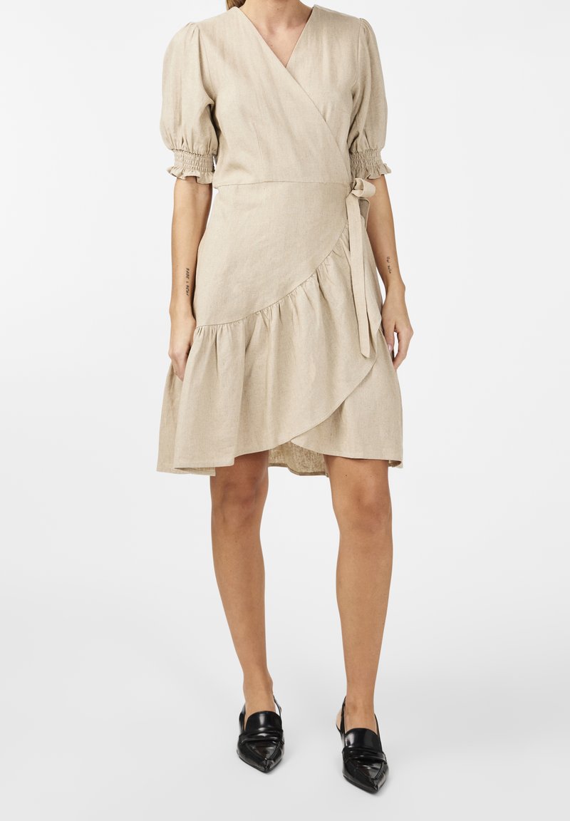 Beige Leinenwickelkleid mit puffärmel, asymmetrischem Saum, raffinierten Akzenten und einem Gürtel an der Taille. Getragen mit schwarzen spitzen Ballerinas.