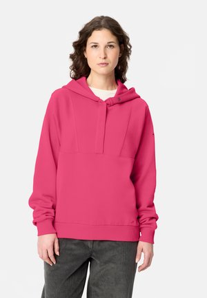 Frau mit lockigem braunem Haar, die einen knallpinken Kapuzenpullover und dunkelgraue Hosen vor einem schlichten hellen Hintergrund trägt.
