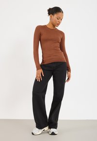 Brūns ribots garām piedurknēm svārku krekls apvienots ar augsta vidukļa melnajiem platajiem džinsiem un pelēkām sporta apavām uz gaiši pelēka fona.