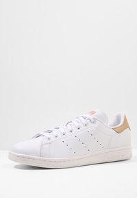 Vit lädersneaker med en slät yta, utrustad med perforerade detaljer på sidan och en tan-färgad accent på hälen. Platta snören, gummisula.
