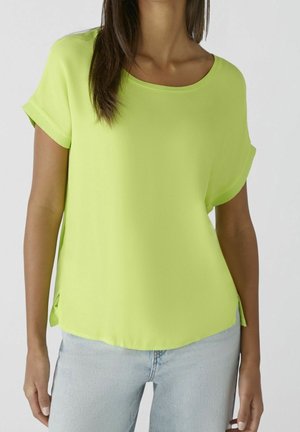 Femme portant un chemisier jaune vif à manches courtes avec un col rond et un jean bleu clair, sur un fond uni.