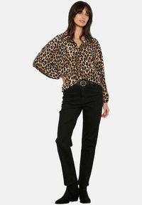 Mujer vestida con una blusa de leopardo de manga larga, pantalones negros de cintura alta con hebilla redonda y botas negras hasta el tobillo, de pie con una mano en la cadera.