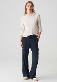 Felpa beige chiara con cappuccio e un motivo testurizzato, abbinata a pantaloni larghi blu navy. Il modello indossa sneakers bianche con suola beige.