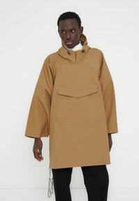 Καφέ αδιάβροχο πουλόβερ με κουκούλα, μεγάλο μπροστινό τσέπη και φαρδιά μανίκια. Διαθέτει κορδόνι στο λαιμό και ματ φινίρισμα.