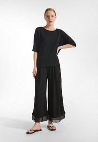 Vrouw met rood haar staat met één hand op haar heup, draagt een los zwart topje, brede zwarte broek met doorschijnende zoom en zwarte sandalen, tegen een witte achtergrond.