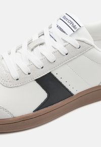 Baskets blanches en cuir et daim avec un accent noir. Elles présentent des lacets plats blancs et une semelle en caoutchouc marron. Le logo sur la languette est visible.