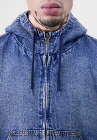 Modrá džínová bunda s kapucí, vybavená zipem na zapínání, šňůrkami a výrazným prošíváním. Texturovaná látka a přední kapsy.