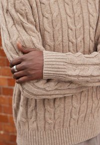 Pull beige en tricot torsadé avec poignets et ourlet côtelés. Texture de tricot épaisse avec un motif de torsade prononcé. Bras croisés, montrant une bague en argent.